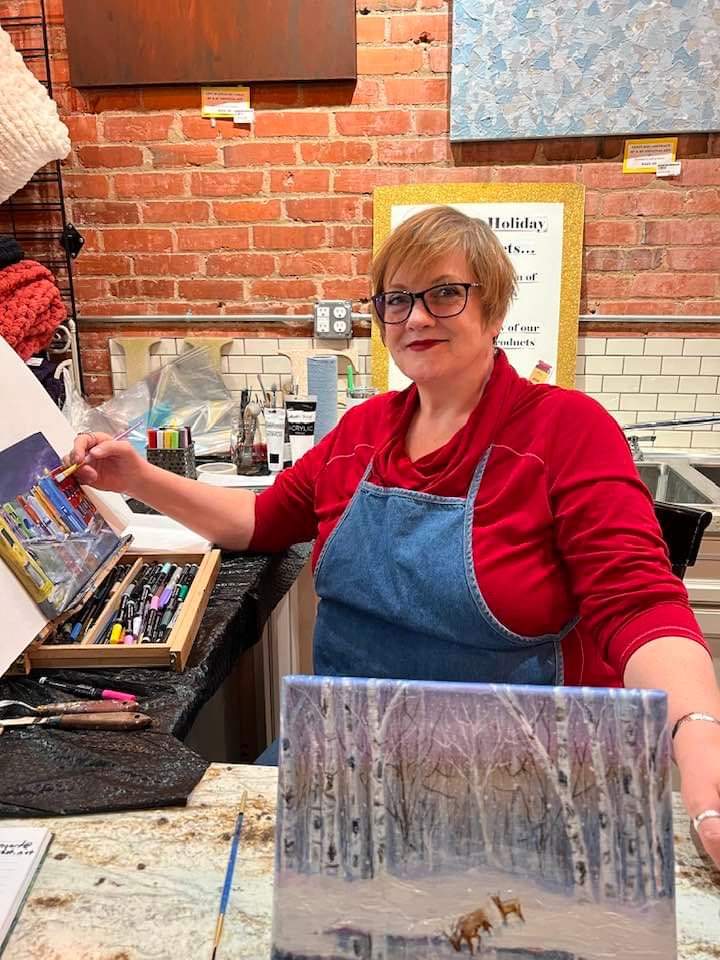 A woman in a red shirt and blue apron sits at a table painting a winter forest scene, with art supplies and a brick wall in the background.