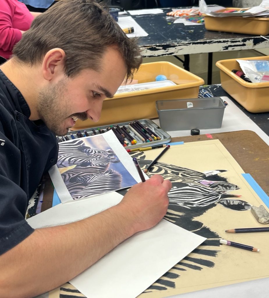 A person draws a detailed zebra illustration in pencil on paper, referencing a printed zebra photo, at a table with art supplies.