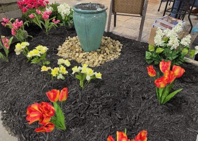 A turquoise ceramic fountain stands on a bed of pebbles, surrounded by artificial flowers and black mulch in a landscaped garden area.