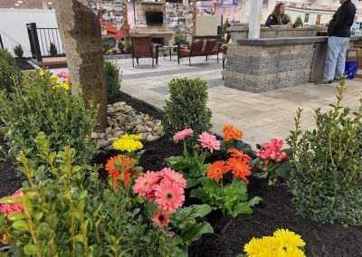 Colorful flowers and green shrubs in a landscaped garden display at an indoor home and garden show, with people talking at a stone counter in the background.
