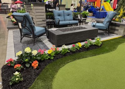 Outdoor patio display with blue cushioned furniture, a rectangular fire pit, and a flower bed bordering artificial turf and mulch.
