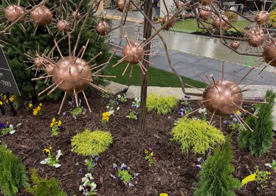 Metallic spherical sculptures with spikes are mounted on branches above a garden bed with mulch, small shrubs, yellow and purple flowers, and a paved area in the background.
