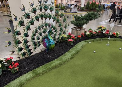 A decorative peacock sculpture stands next to a mini golf putting green, surrounded by flowers and artificial grass inside an indoor exhibition space.