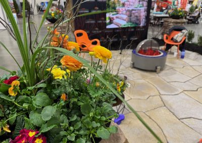 A close-up of colorful flowers in the foreground with a patio display, orange chairs, a fire pit, and a screen showing a garden scene in the background at an indoor event.