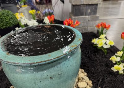 A close-up of a blue ceramic water fountain surrounded by white, yellow, and red flowers with a stone path and indoor event setting in the background.