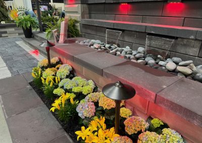 A landscaped garden features yellow lilies, hydrangeas, modern stonework, outdoor lamps, a rock-filled water feature, and red accent lights.