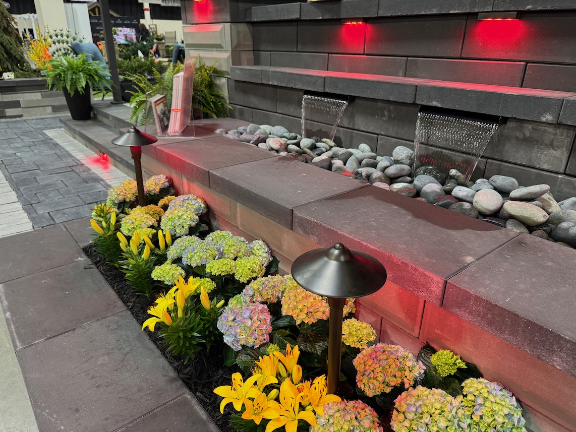A landscaped garden features yellow lilies, hydrangeas, modern stonework, outdoor lamps, a rock-filled water feature, and red accent lights.