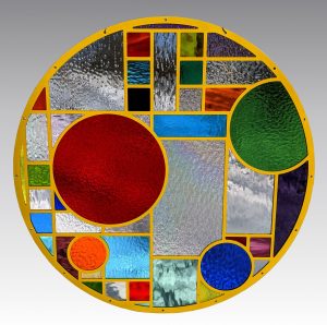 A round stained glass window with yellow framing, featuring various textured and colored glass sections in red, green, blue, and clear patterns arranged in geometric shapes.