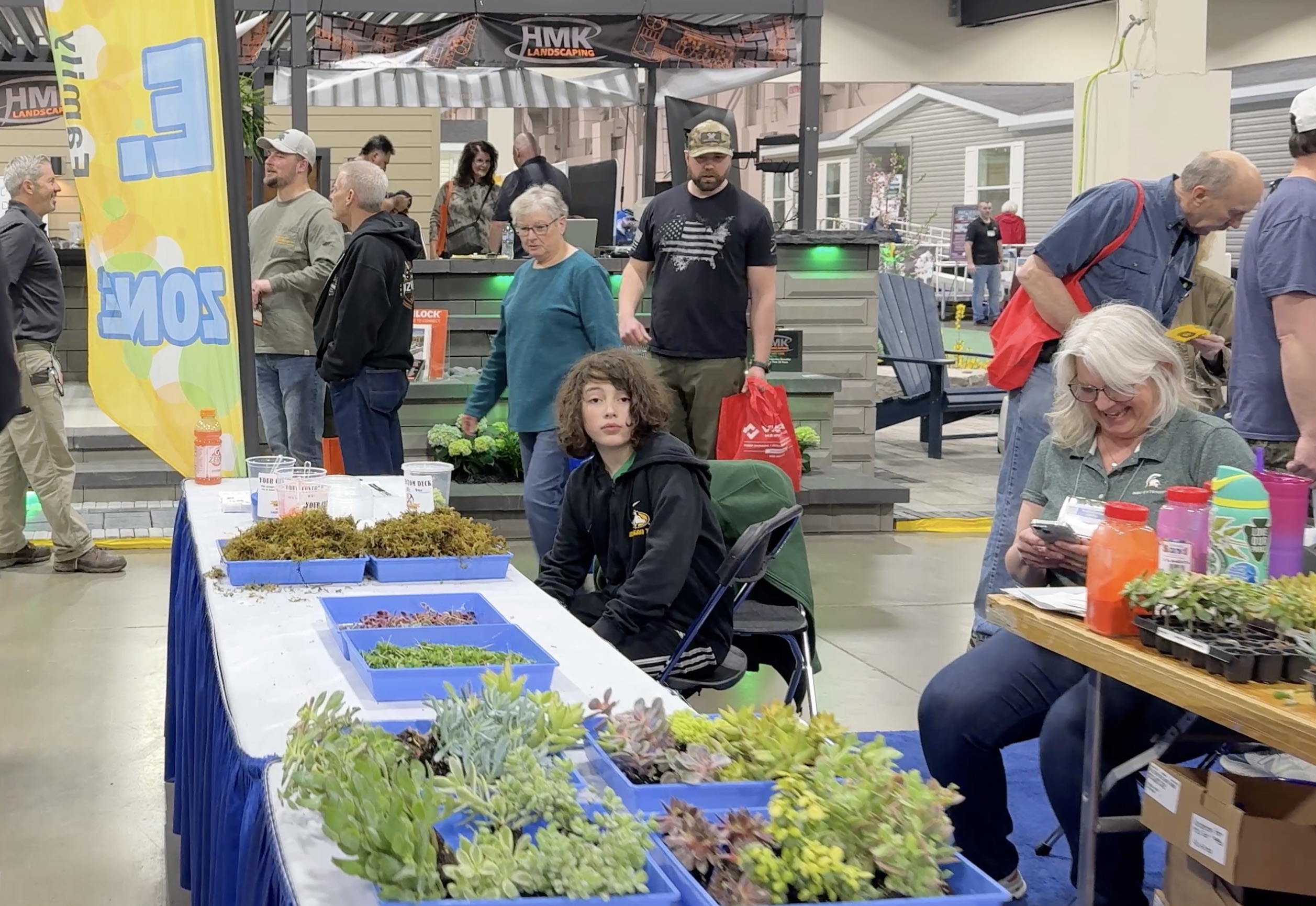 Screenshot People are gathered at an indoor event with tables displaying various potted succulents. Some are sitting behind the table, while others walk around or view the plants.